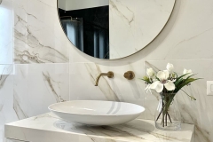 Erretre Ceramiche realizzazione di un bagno moderno in una villetta a Magenta, con piano lavabo di colore chiaro, lavabo ovale di colore bianco con rubinetti e pomello di colore dorato, e, uno specchio in vetro di forma rotonda alla parete