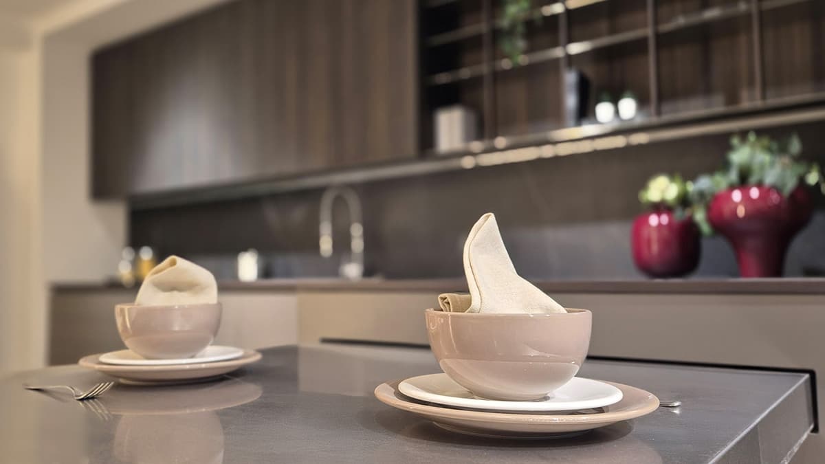 Dettaglio Penisola e allestimento tavola minimalista della Cucina Elmar Modello Slim. Vista sui pensili in legno scuro e vasi decorativi rossi.